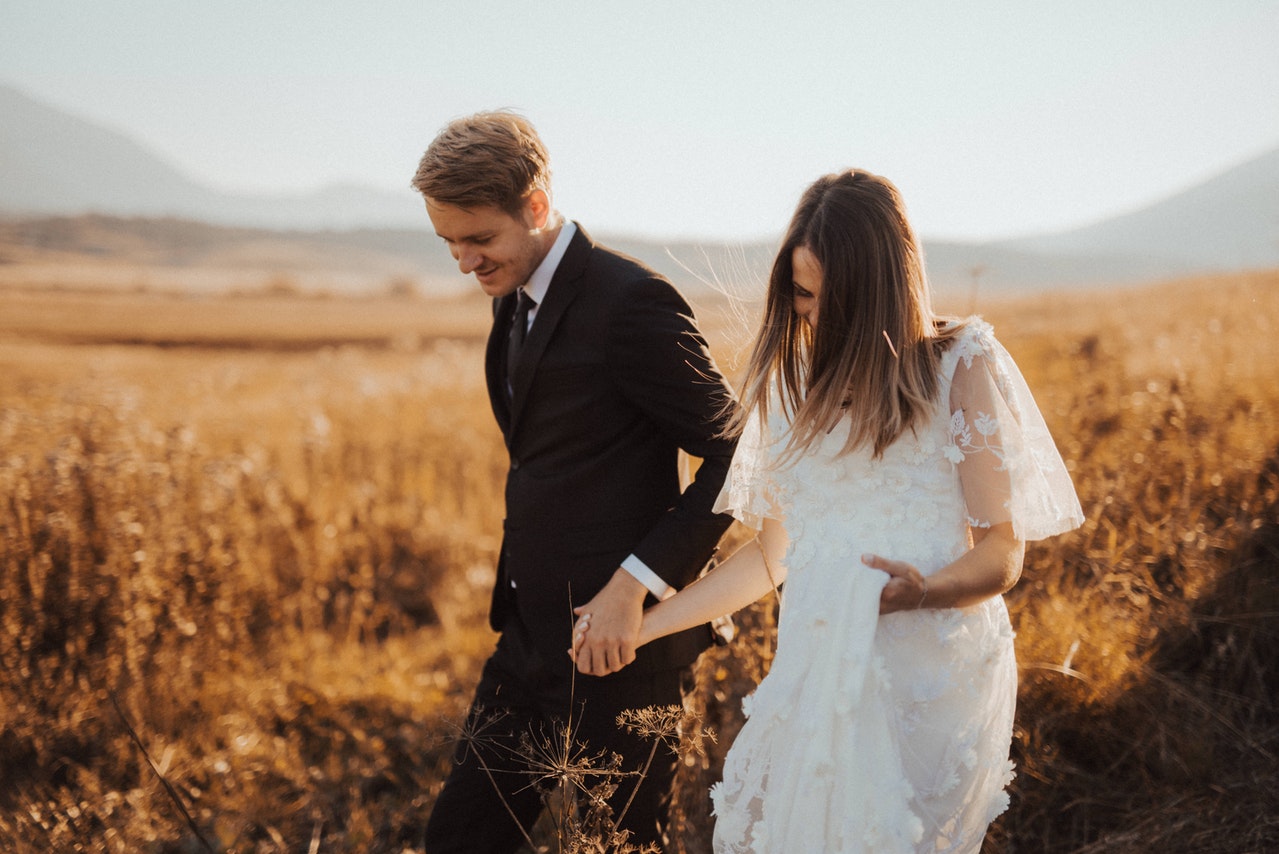 Photo of Man in Black Formal Suit Holding Woman's Hand in White Dress walking through tall dried grass